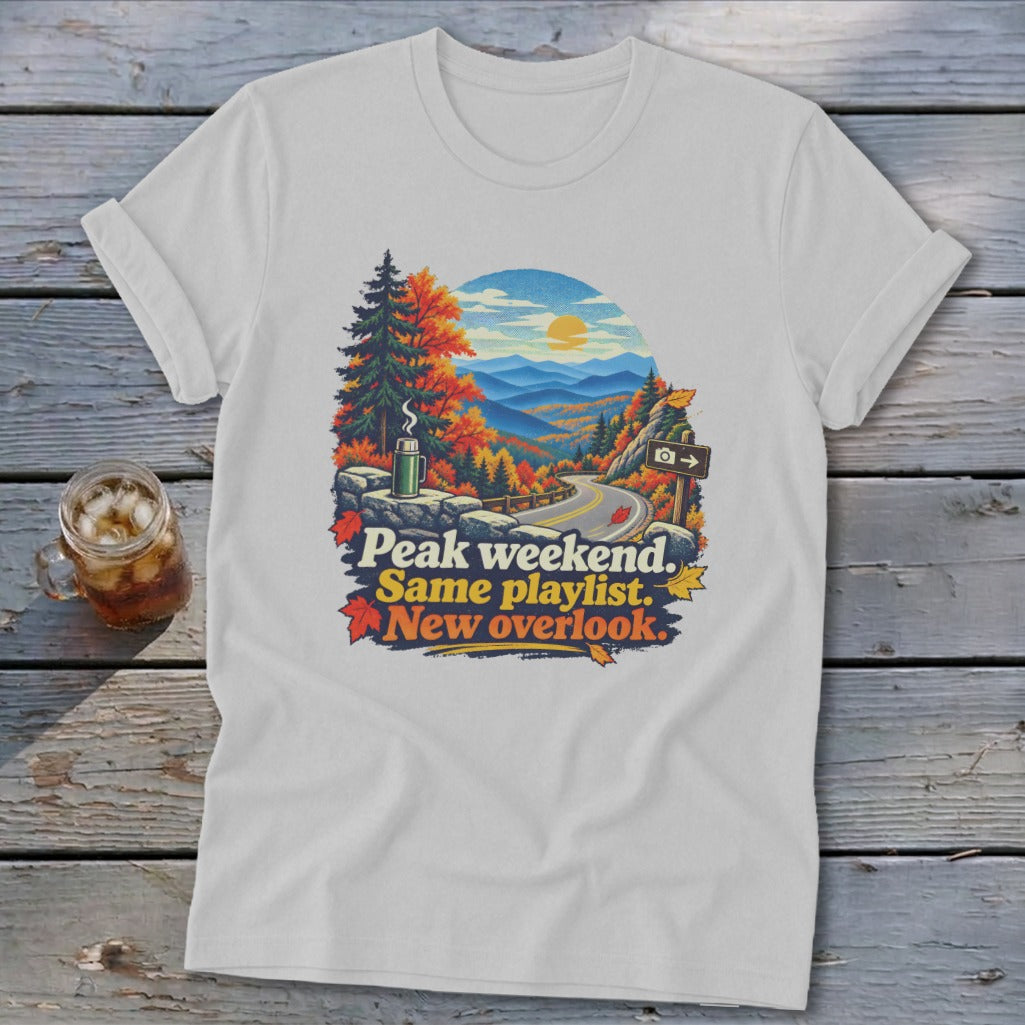 White t-shirt with mountain landscape graphic and text "Peak weekend. Same playlist. New overlook."