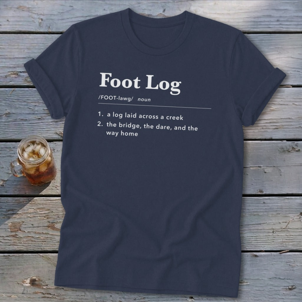 A dark blue t-shirt with white dictionary-style text defining Foot Log lies on a wooden surface beside a glass of iced tea.