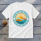 A white t-shirt featuring a circular graphic with mountains, a rising sun, and text reading BORN IN THE HOLLER RAISED ON THE RIDGE.