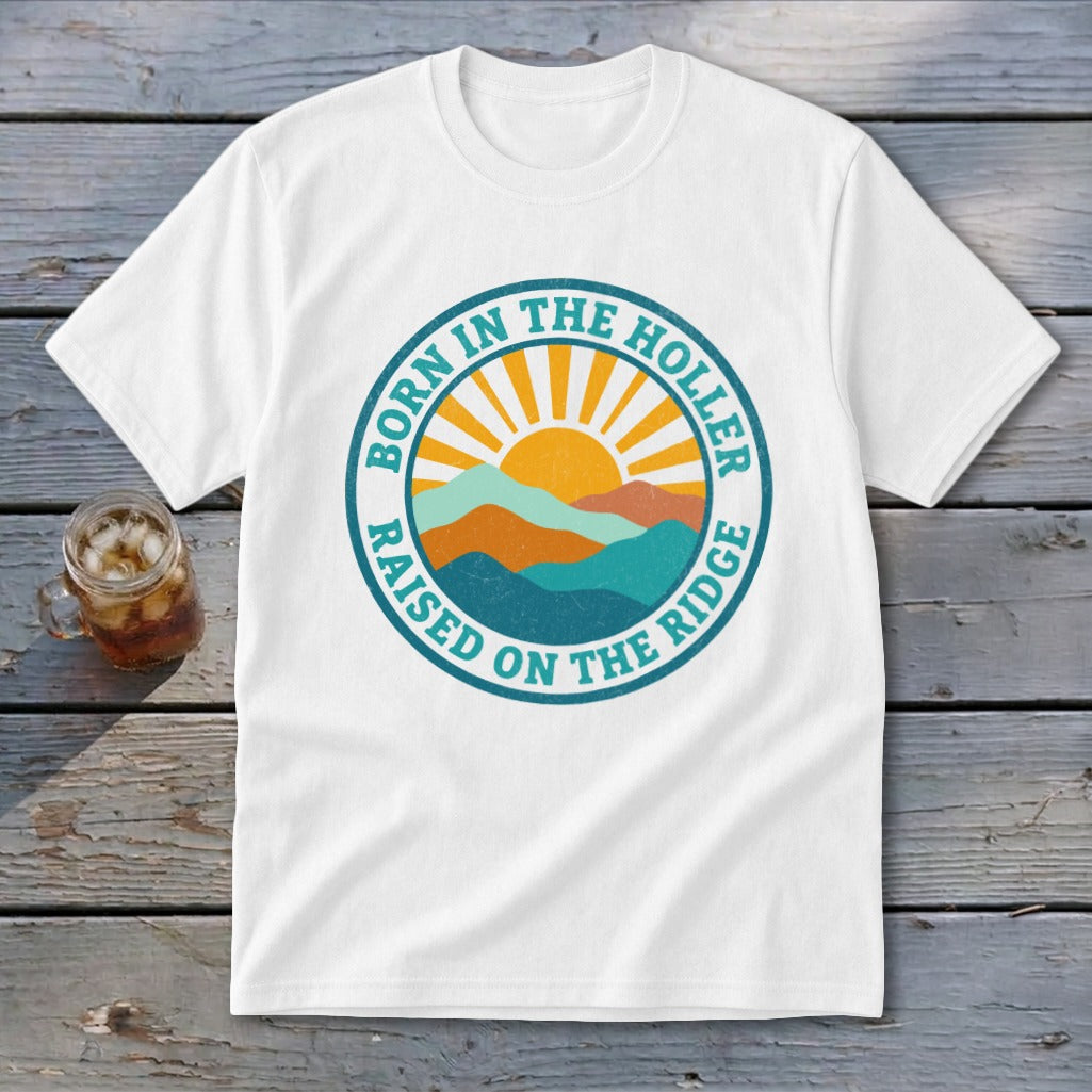 A white t-shirt featuring a circular graphic with mountains, a rising sun, and text reading BORN IN THE HOLLER RAISED ON THE RIDGE.