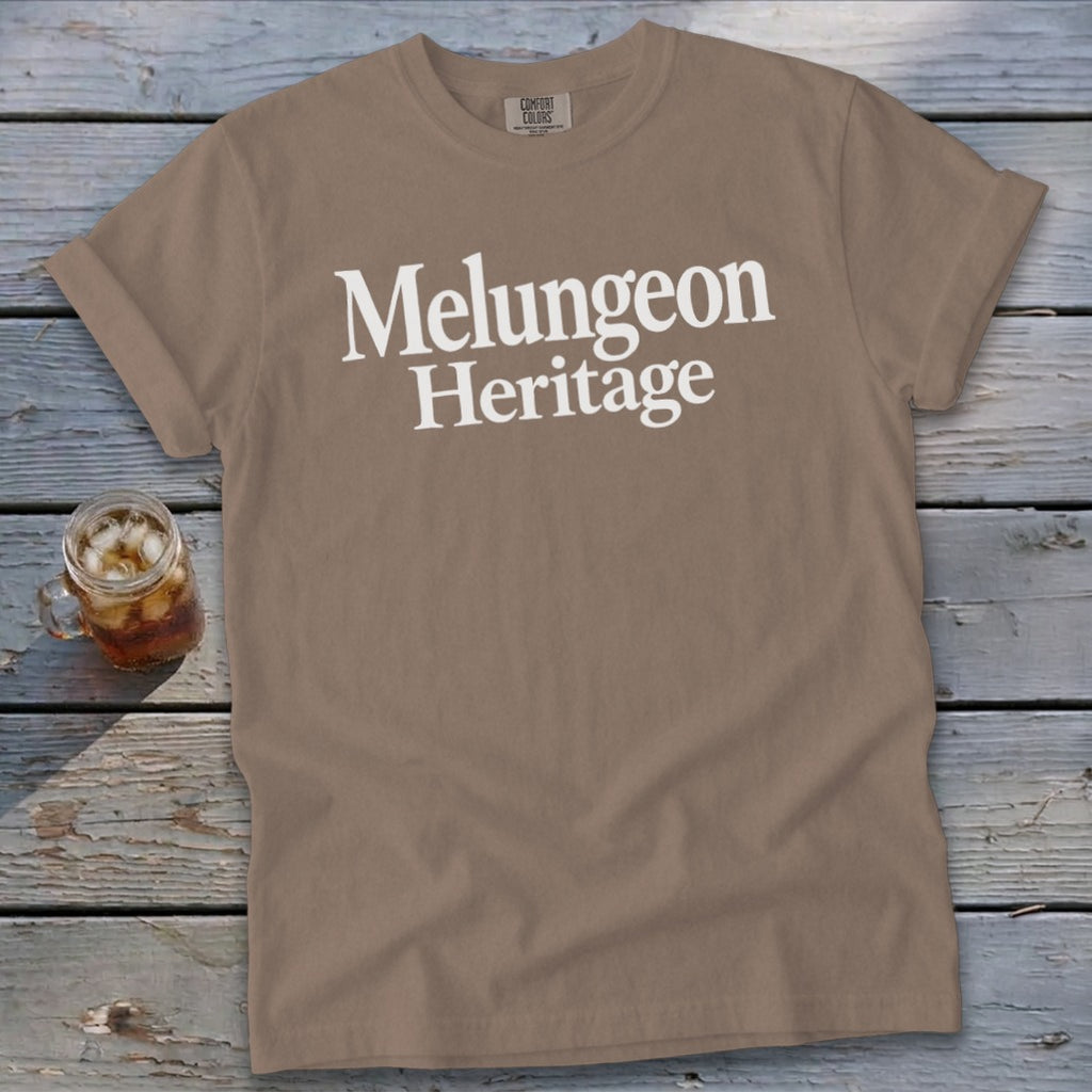 Brown crewneck t-shirt with white Melungeon Heritage text displayed across the chest, resting on wood planks.
