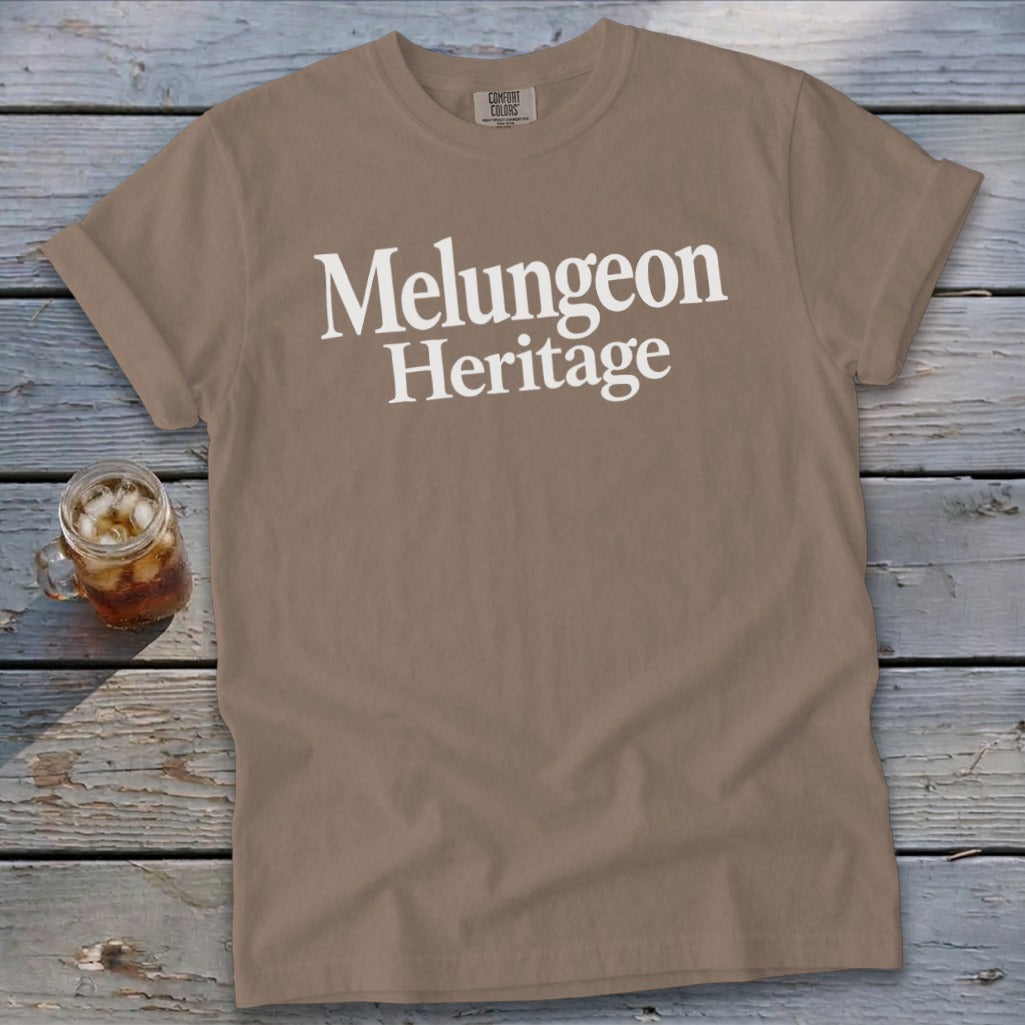 Brown crewneck t-shirt with white Melungeon Heritage text displayed across the chest, resting on wood planks.