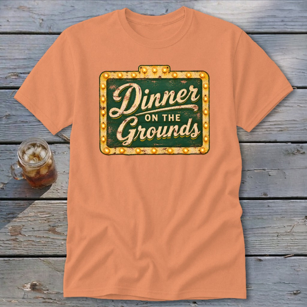 Peach-colored short-sleeve t-shirt featuring a green sign graphic with the text Dinner on the Grounds.