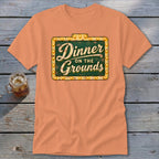 Peach-colored short-sleeve t-shirt featuring a green sign graphic with the text Dinner on the Grounds.