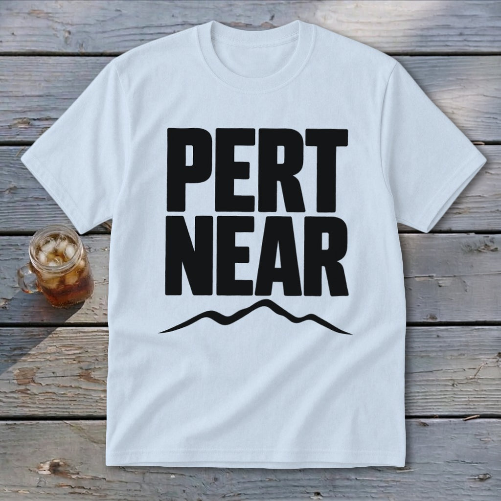 A white t-shirt laid flat on a wooden surface, featuring the black text PERT NEAR above a mountain graphic.