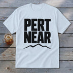 A white t-shirt laid flat on a wooden surface, featuring the black text PERT NEAR above a mountain graphic.