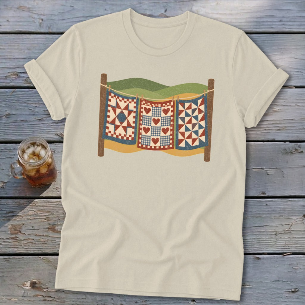 A cream-colored t-shirt displays a vintage-style graphic of three colorful quilts hanging on a clothesline against a stylized landscape.