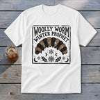 A white t-shirt featuring a black and brown block print design of a woolly worm and text reading WOLLY WORM WINTER PROPHET.