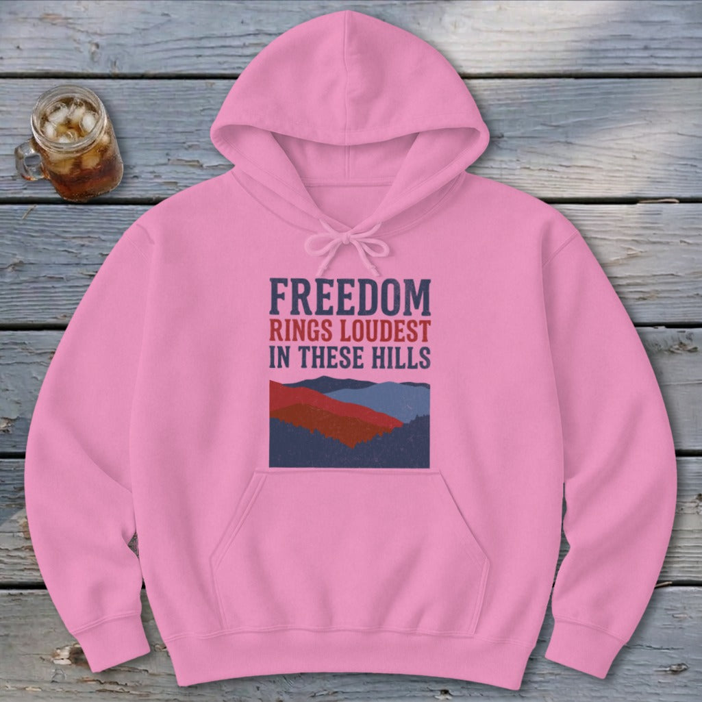 A pink hooded sweatshirt featuring the text "FREEDOM RINGS LOUDEST IN THESE HILLS" above a stylized mountain graphic, lying on a wooden surface with a glass of iced tea.