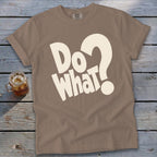 Taupe crewneck t-shirt with rolled sleeves featuring bold, cream-colored text reading Do What? lying on a weathered wooden surface.