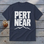 Navy blue short-sleeve t-shirt with white text reading PERT NEAR above a mountain range graphic, laid on weathered wooden planks.