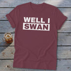 A maroon crewneck t-shirt with rolled sleeves lies flat on weathered wooden boards beside a glass of iced tea.
