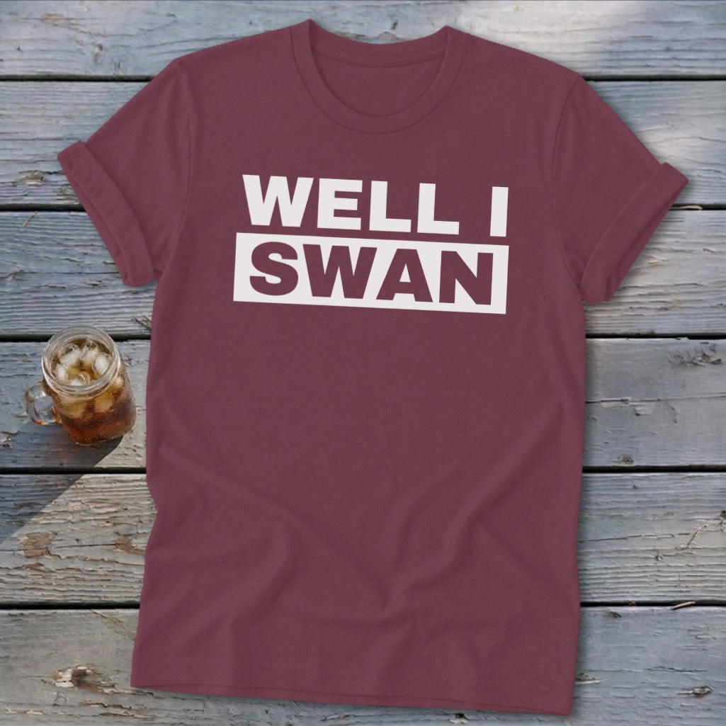 A maroon crewneck t-shirt with rolled sleeves lies flat on weathered wooden boards beside a glass of iced tea.