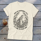 A cream-colored t-shirt with a black vintage illustration of a bird, corn stalks, and text stating WHEN THE WHIPPOORWILL CALLS IT'S TIME TO PLANT CORN.