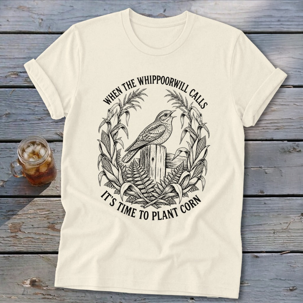 A cream-colored t-shirt with a black vintage illustration of a bird, corn stalks, and text stating WHEN THE WHIPPOORWILL CALLS IT'S TIME TO PLANT CORN.