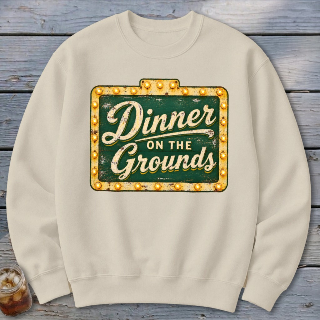 Cream-colored crewneck sweatshirt featuring a green and gold marquee sign graphic with the text Dinner on the Grounds.
