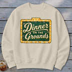 Cream-colored crewneck sweatshirt featuring a green and gold marquee sign graphic with the text Dinner on the Grounds.