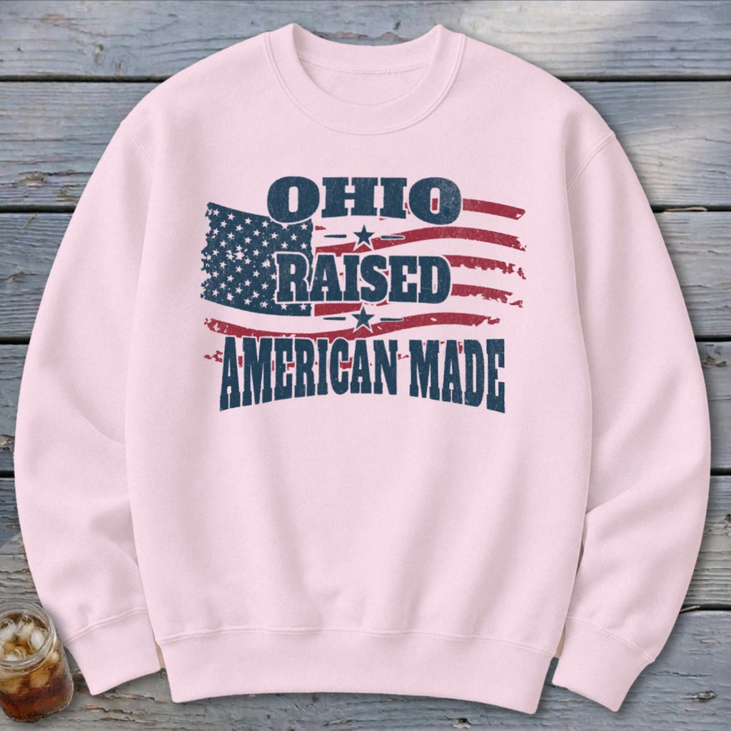 A light pink crewneck sweatshirt featuring a distressed American flag graphic with text reading OHIO RAISED AMERICAN MADE.