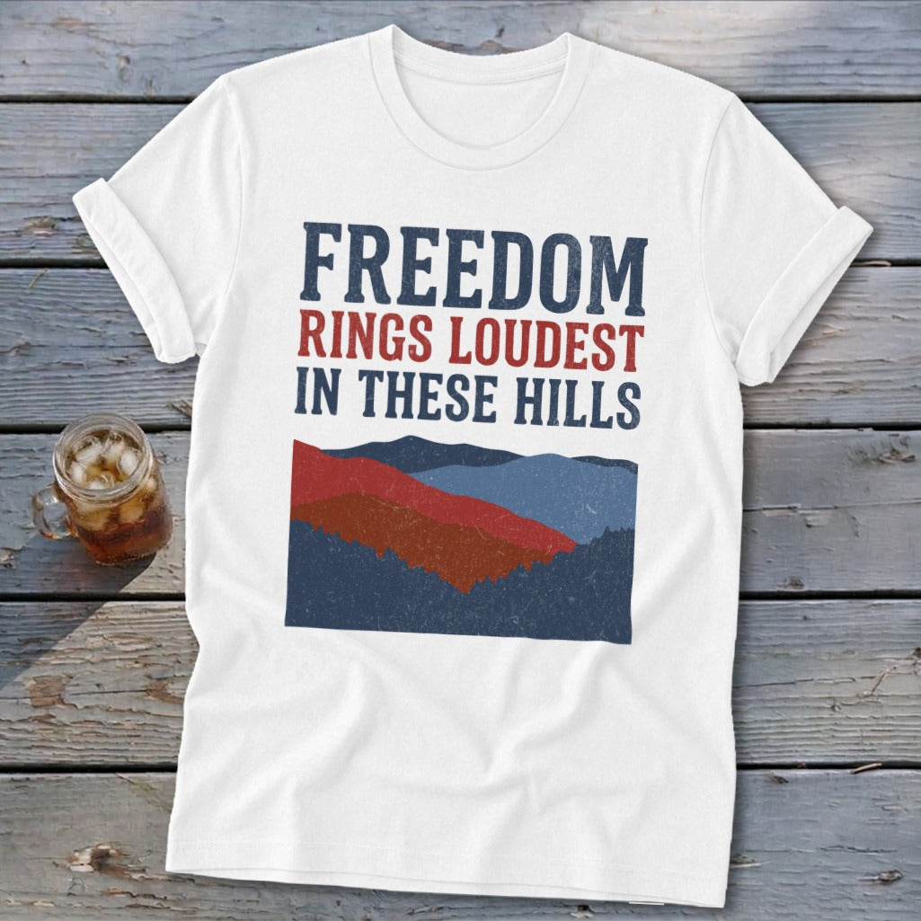 White t-shirt featuring distressed blue and red text reading Freedom Rings Loudest In These Hills above a stylized mountain graphic.