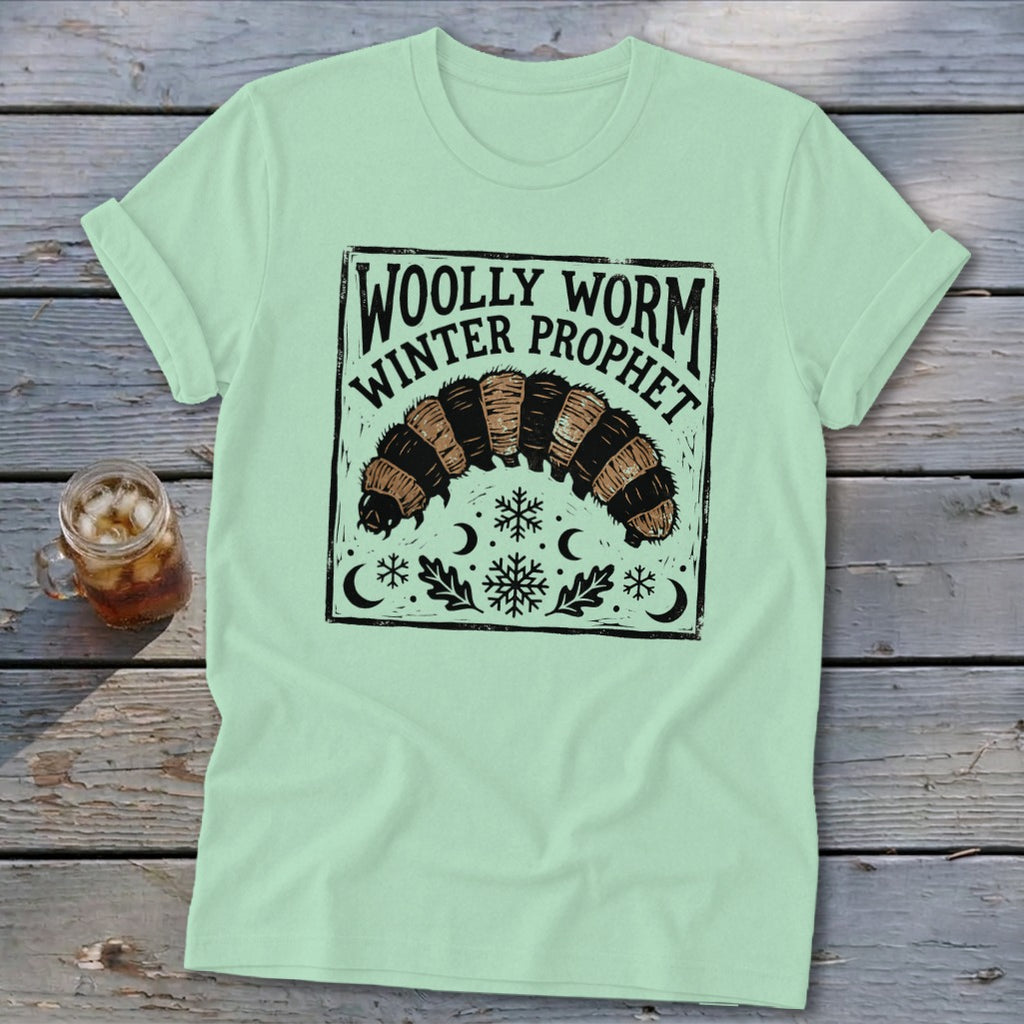 Light green t-shirt featuring a graphic of a woolly worm caterpillar, snowflakes, moons, and text reading Woolly Worm Winter Prophet.