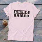 Pink t-shirt with black text "CREEK RAISED" and a drink.