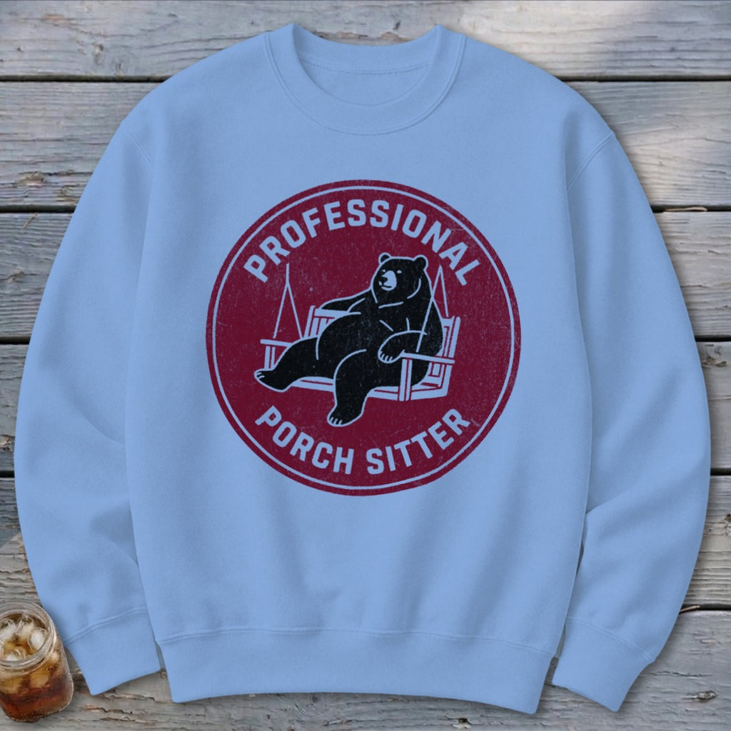 A light blue crewneck sweatshirt featuring a circular red graphic with a bear relaxing on a porch swing and text reading PROFESSIONAL PORCH SITTER.