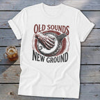A white t-shirt features a graphic print of a hand playing a banjo and text that reads Old Sounds New Ground.