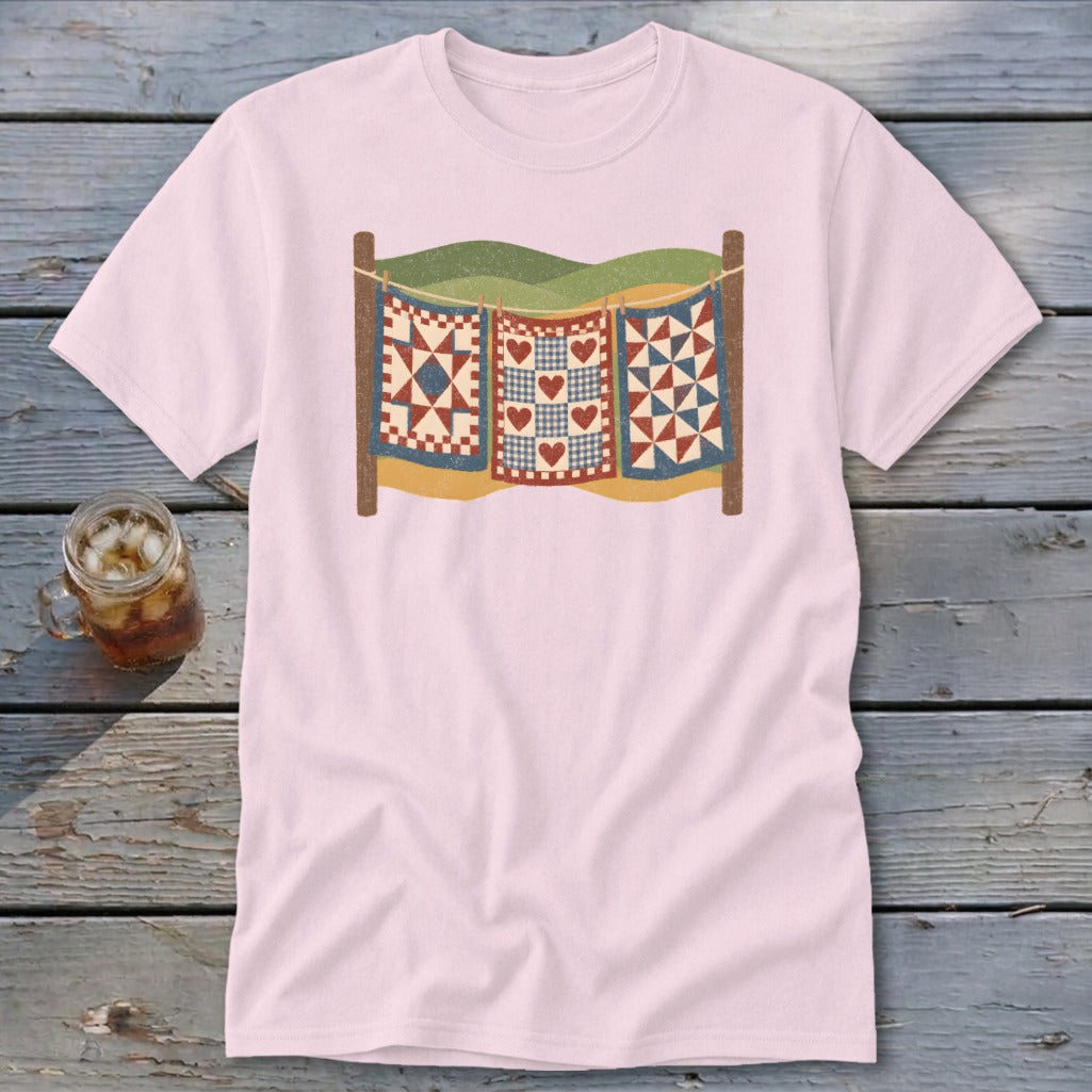 A light pink t-shirt featuring a retro graphic of three quilts hanging on a clothesline.