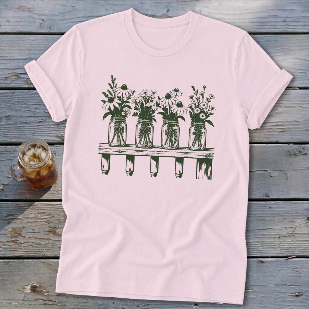 Light pink short-sleeve t-shirt featuring a green graphic of four mason jars filled with wildflowers resting on a wooden shelf.