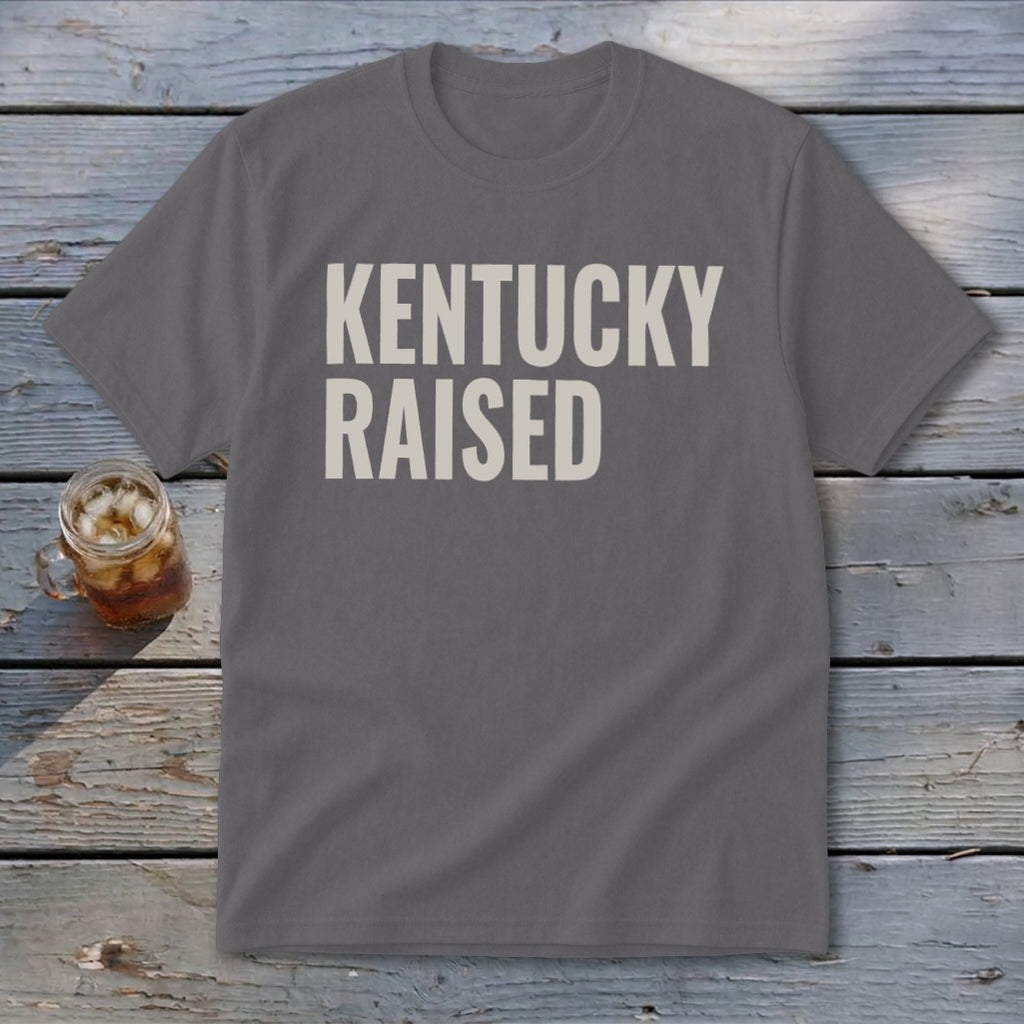 Grey t-shirt with "KENTUCKY RAISED" text in off-white on a wooden background.