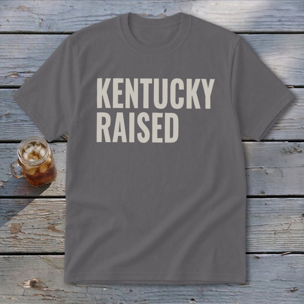 Grey t-shirt with "KENTUCKY RAISED" text in off-white on a wooden background.