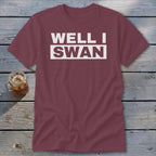 Maroon short-sleeved t-shirt with white text reading WELL I SWAN laid on a rustic wooden table next to a glass of iced tea.