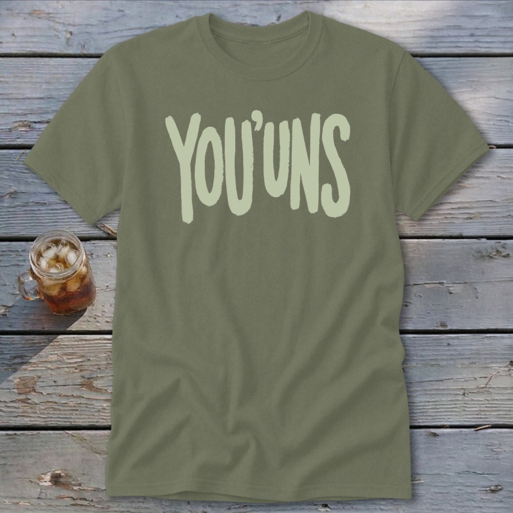 Olive green t-shirt with "YOU'UNS" text and a drink on a wooden surface.