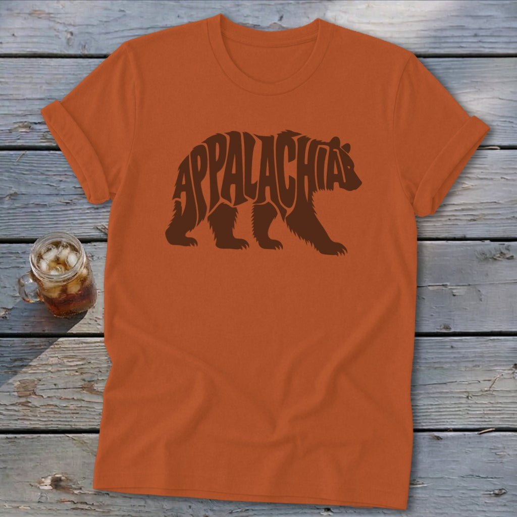 An orange t-shirt with a brown bear graphic made of the word Appalachia rests on a wooden surface.
