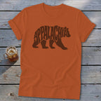 An orange t-shirt with a brown bear graphic made of the word Appalachia rests on a wooden surface.