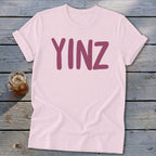 Pink t-shirt with "YINZ" in dark pink text, beside a glass of iced tea.