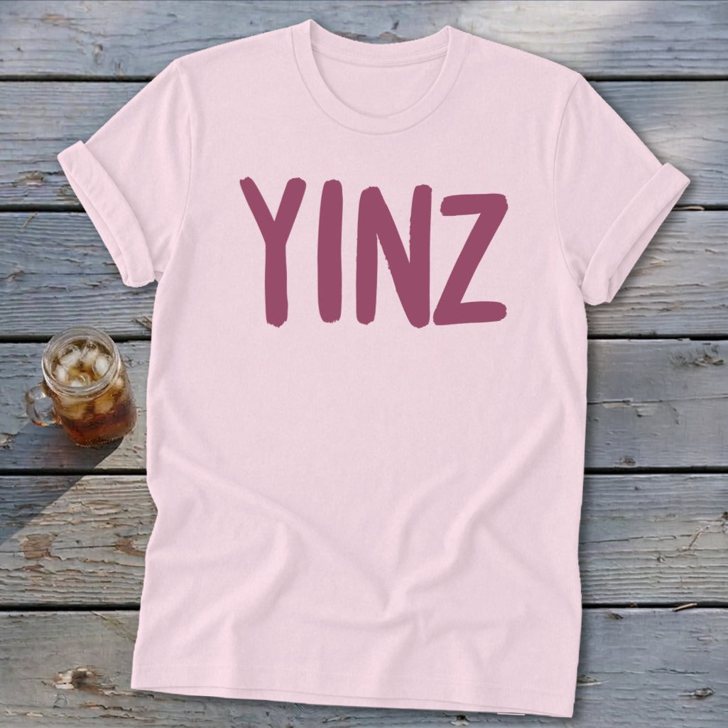 Pink t-shirt with "YINZ" in dark pink text, beside a glass of iced tea.