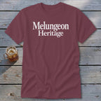 Maroon short-sleeved t-shirt with white text reading "Melungeon Heritage" centered on the chest, laid flat on a rustic wooden surface next to a glass of iced tea.