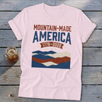 A pink t-shirt featuring a red, white, and blue graphic with the text MOUNTAIN-MADE AMERICA 1776-2026.