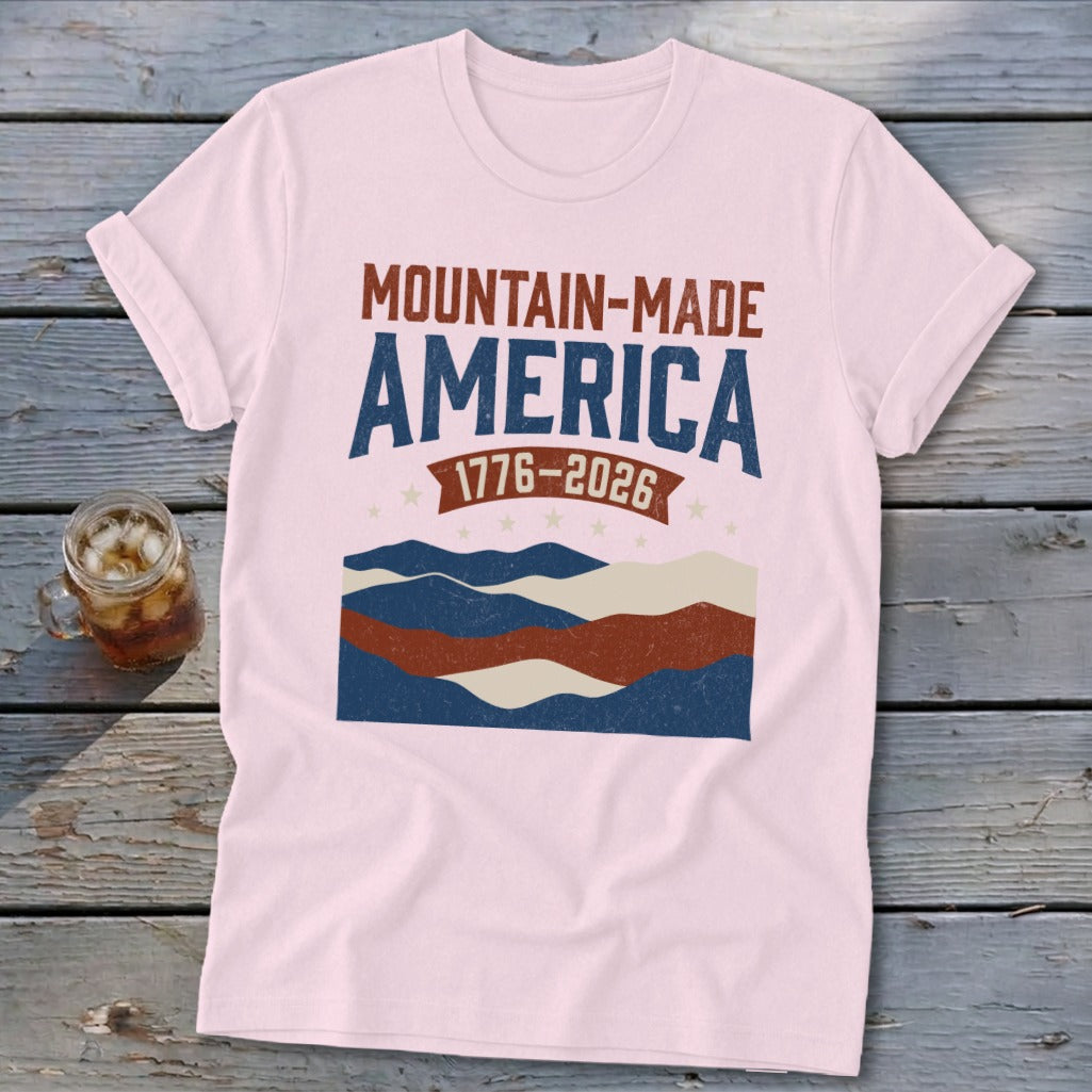 A pink t-shirt featuring a red, white, and blue graphic with the text MOUNTAIN-MADE AMERICA 1776-2026.