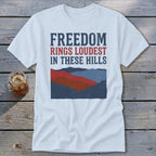 A white t-shirt featuring the text "FREEDOM RINGS LOUDEST IN THESE HILLS" above a mountain graphic.