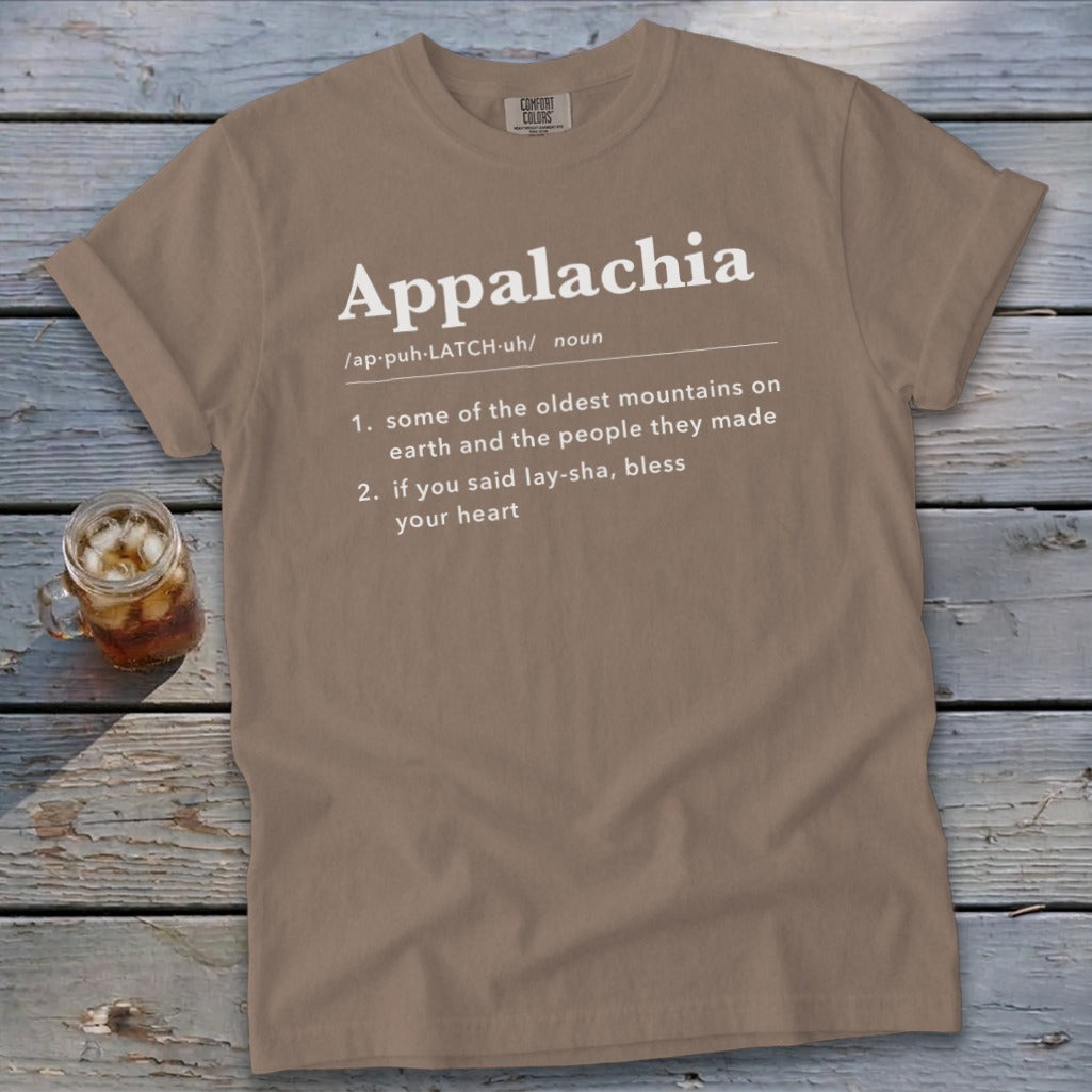 Brown t-shirt featuring dictionary-style text defining Appalachia, laid flat on a rustic wooden surface next to a glass of iced tea.